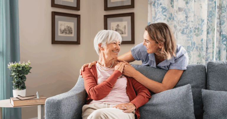 Smiling caregiver with hand over the shoulder of a happy elderly woman sitting on the couch, representing the results of quality home care services.