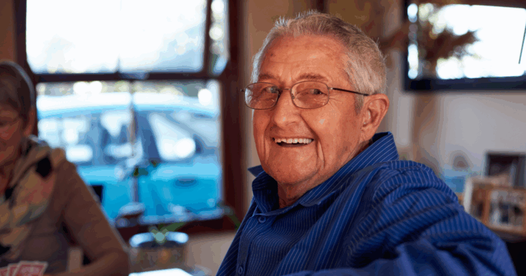 A happy, smiling senior man is sitting at a table and looking over his shoulder, enjoying aging in place as a result of successful of long-distance caregiving.